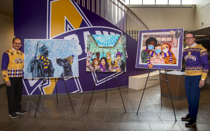 Two men in purple and yellow shirts patterned with snowflakes stand on either side of three easels holding large images of winter holiday cards 