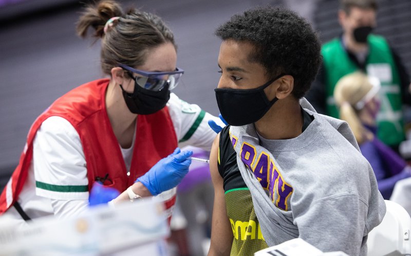 A student wearing a UAlbany shirt receives a COVID-19 vaccine from a volunteer, wearing a red vest. Both are wearing black masks.