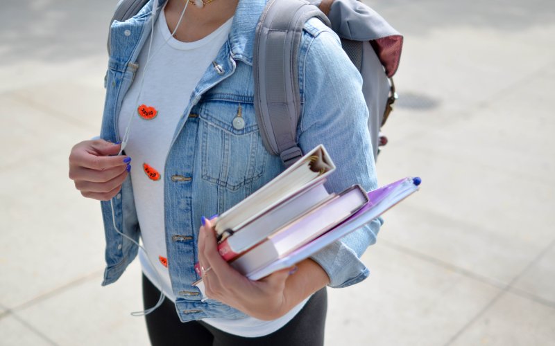 A girl holds three books and a notebook, and is wearing a backpack.