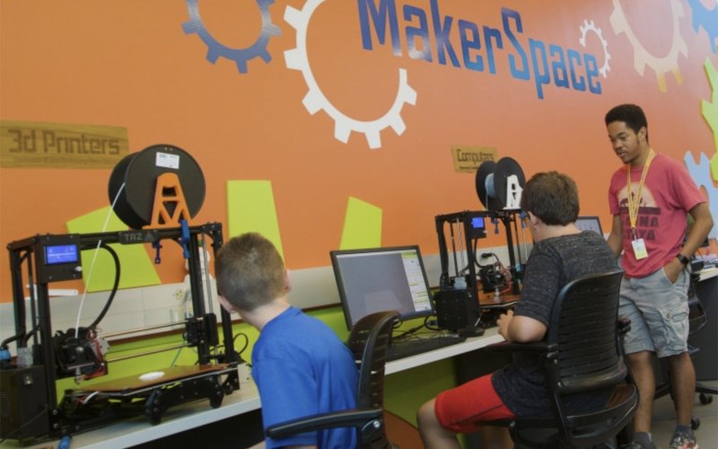 Two children sit at a table with computers and 3D printers are helped by a young man indoors, with a sign MakerSpace on the wall.