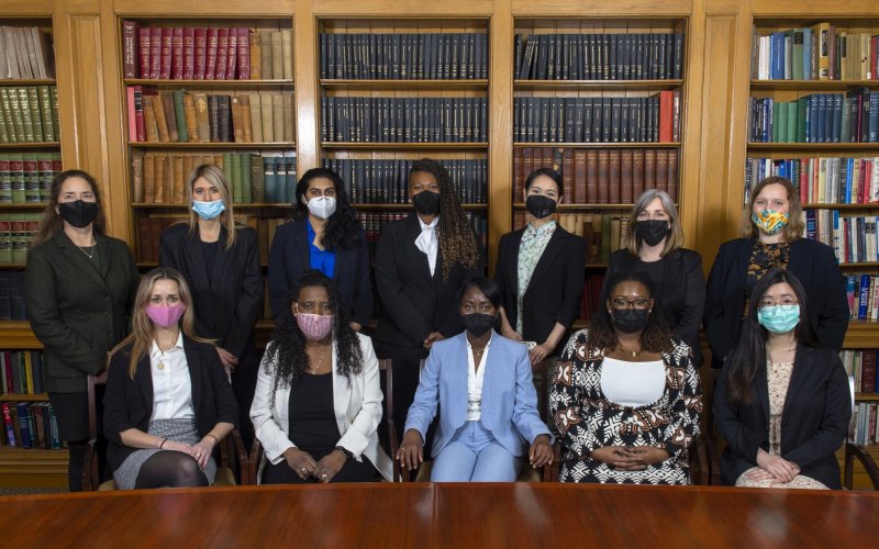 Seven women stand behind five seated women, all wearing facemasks and professional office clothing, in front of shelves of books.