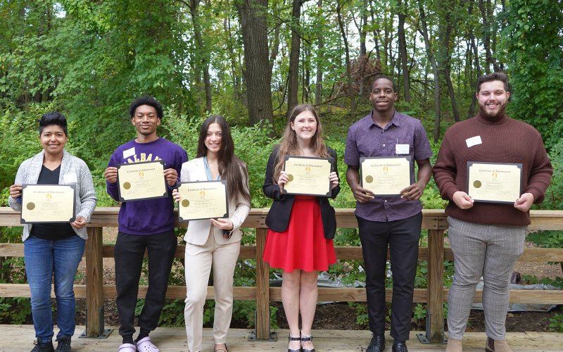 Six smiling students stand in a row in front of a wooden fence, each holding a scholarship certificate
