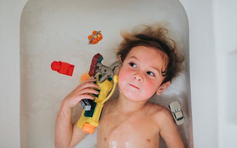 A young boy playing in a bathtub, holding one toy and surrounded by others.