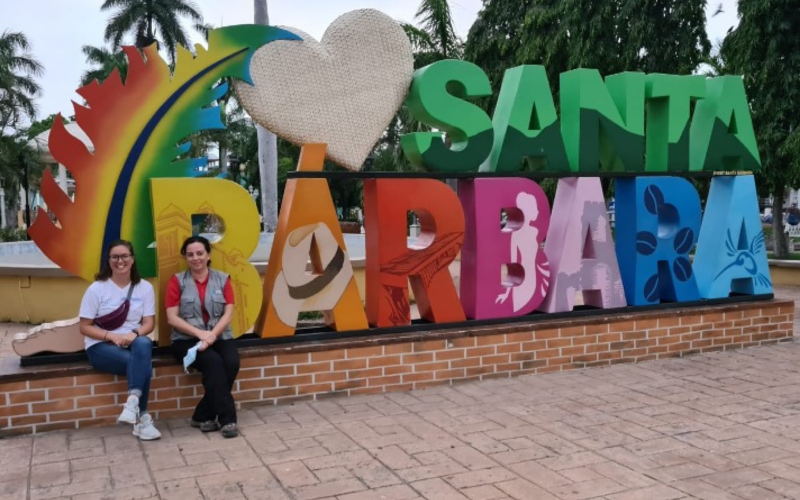 Aubrey Racz and a colleague sit on a brick wall in front of a colourful sign that says "Santa Barbara".