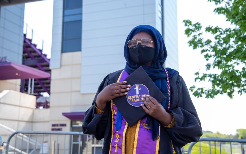 A soman wearing a headscarf, face mask and glasses along with graduation robes holds a circular sticker that says 1st generation graduate.