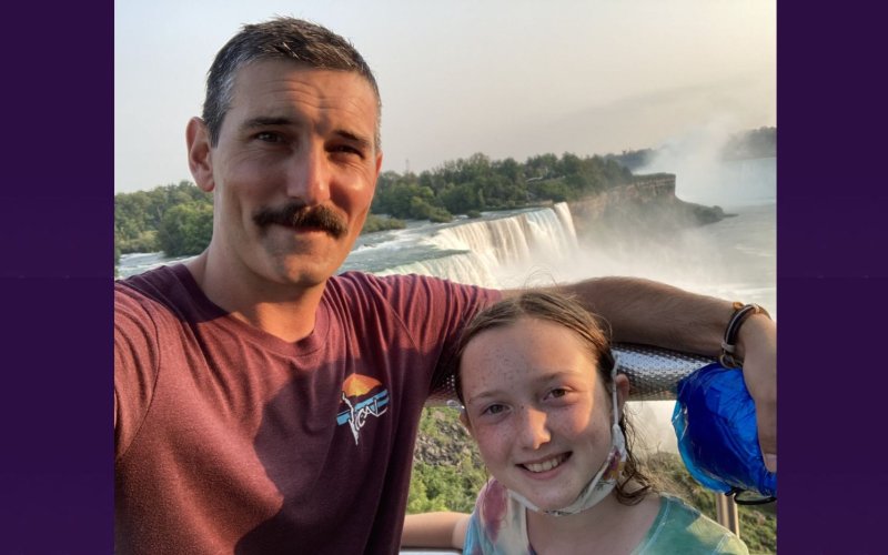 A man with a mustache and his freckle-faced young daughter smile while standing together in front of Niagara Falls.