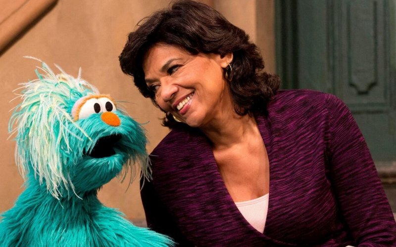 Sonia Manzano, in a purple sweater, leans in and smiles at a large, fuzzy, light blue Muppet
