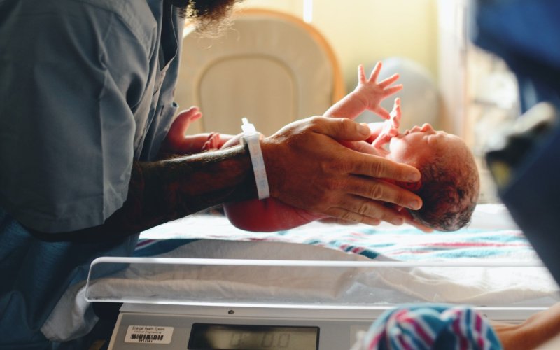 A person in gray medical scrubs and a hospital writband places a newborn infant on an electronic scale