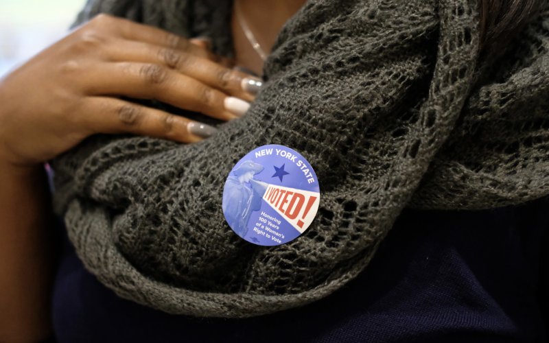 An African American woman  shows of her New York State I Voted sticker on her sweater.
