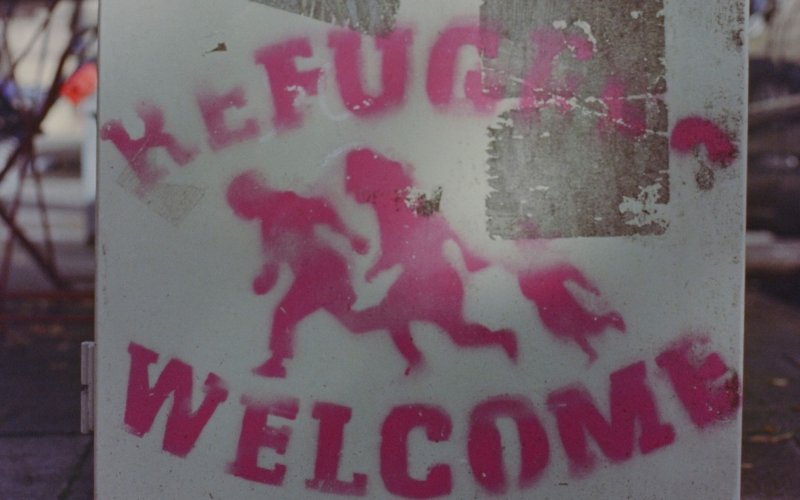 a sign stencilled in pink spray paint shows three people running and the word "refugees welcome"