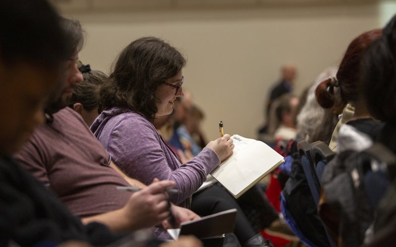 A young woman takes notes seated amongst other people in an audience as poet Sharon  Olds speaks during a New York State Writers Institute craft conversation in the UAlbany Campus Center Multipurpose Room on Thursday, October 24, 2019. (photo by Patrick Dodson)