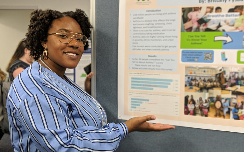 A student stands in front of her poster, holding her hand toward it inviting you to view her work.