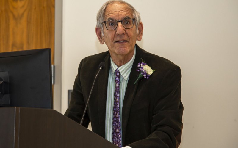 Professor Berman at a podium, wearing glasses, a brown jacket and purple tie, and a flower in his lapel.