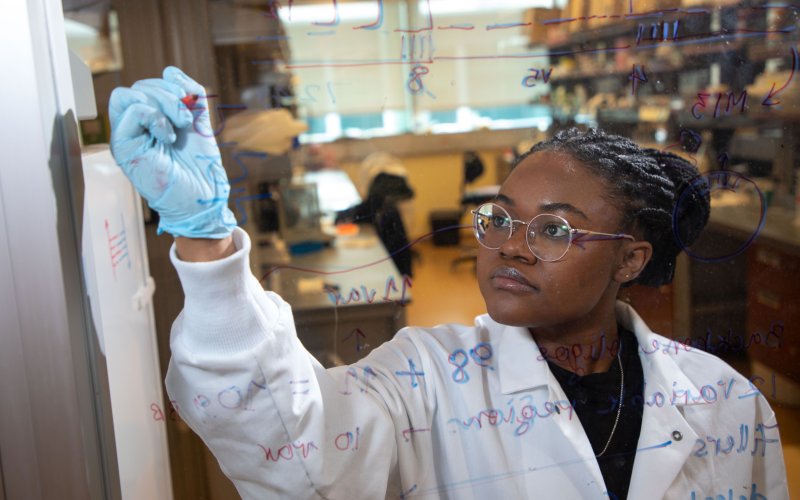 A woman in a lab coat and blue goves is seen behind a pane of glass she is writing formulas on.