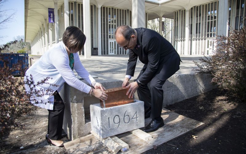 A man and a woman place a metal box into a cement cornerstone with 1964 engraved on it