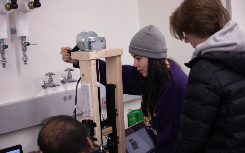 Engineering students at UAlbany work indoors on a rudimentary tensiometer device made of wood with a metallic gear at the top.