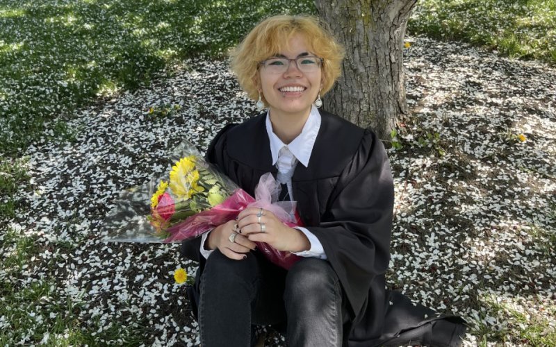 Maya Lee '22 in glasses holds a bouquet of flowers and offers a big smile while sitting under a tree
