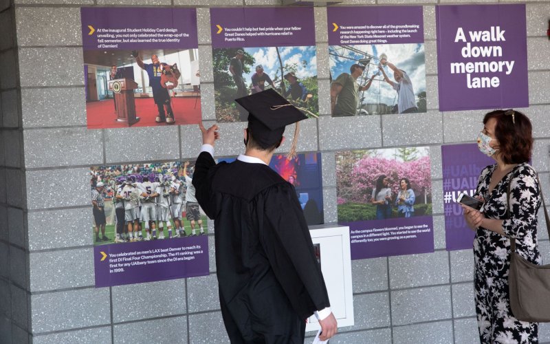 A student in graduation regalia points to photos on a wall while standing with a woman.
