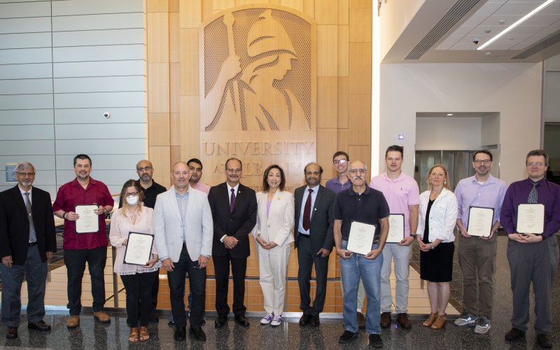 A group of faculty stand in front of the UAlbany Minerva logo in the ETEC building with recognition awards in hand.