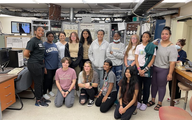 A group of female students sit and stand in front of physics equipment in a lab at UAlbany.