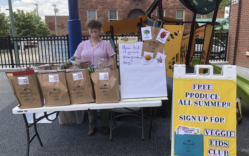 Melissa Toback stands outside behind a table that holds several large brown bags filled with produce.