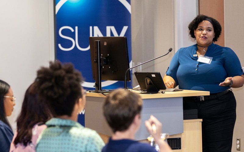 A woman speaks at a podium indoors in front of a seated crowd with a SUNY logo behind her.
