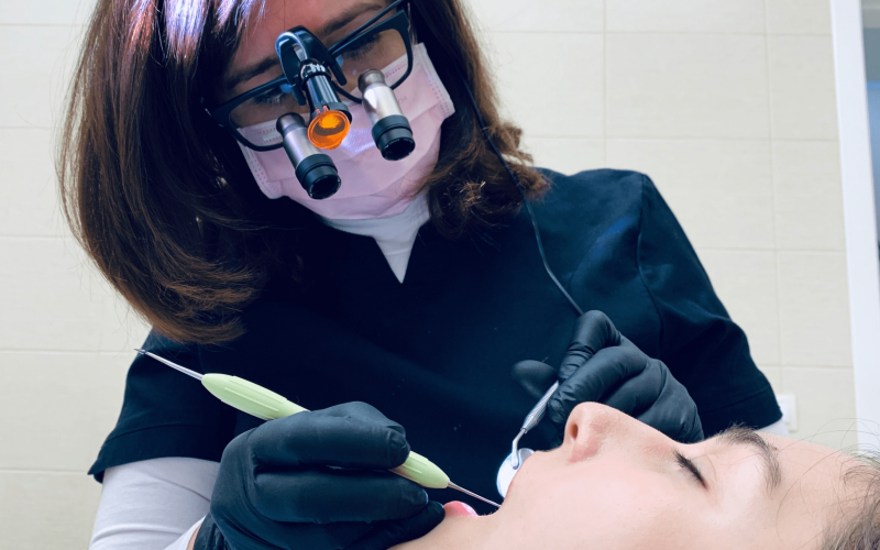 Image of a dental cleaning: A woman with dark brown hair wearing dark-rimmed glasses with magnifying attachments, wearing a paper face mask and latex gloves, cleans the teeth of a reclining person in the foreground using metal dental tools. 