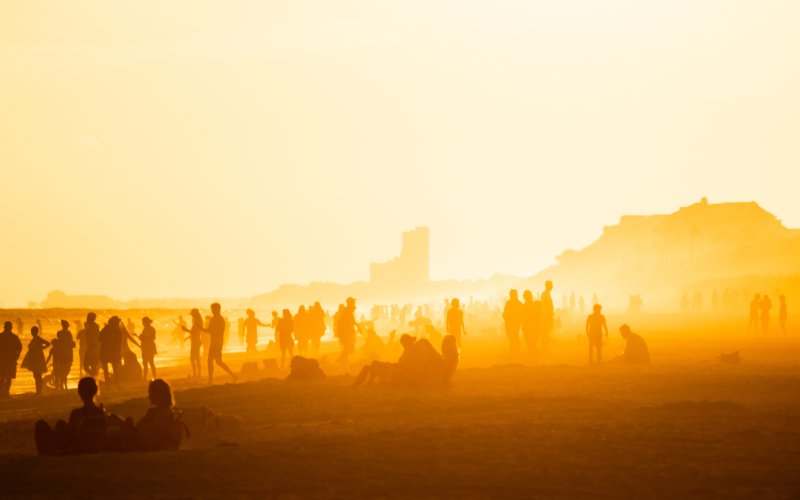Beach crowded with people at sunset. The image is rich in orange and red tones; people are primarily cast as silhouettes. 