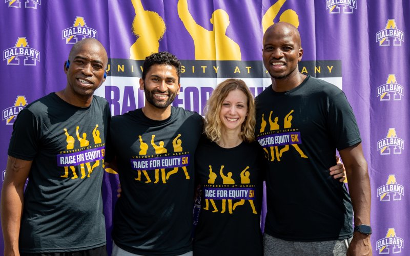 Four smiling adults wearing black "Race for Equity 5k" T-shirts stand in front of a purple fabric backdrop printed with University at Albany logos.