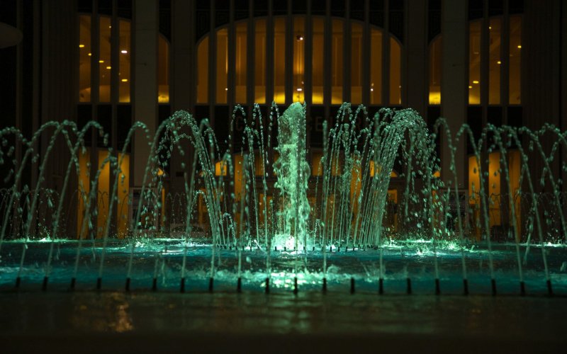 A fountain is lit with green lights, and seen in front of a building with glowing yellow lights from its windows