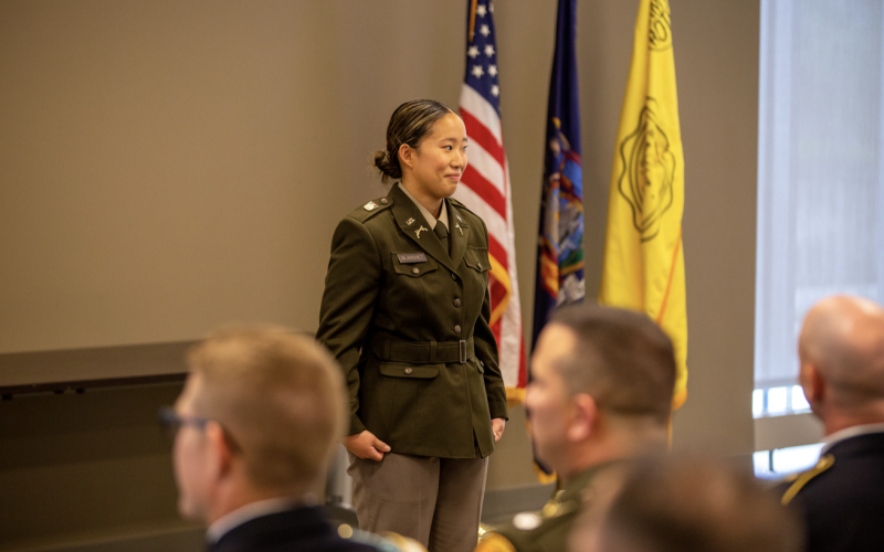A young woman in a U.S. Army uniform stands at attention in front of the U.S., Army and N.Y. State flags indoors with other officers in attendance.
