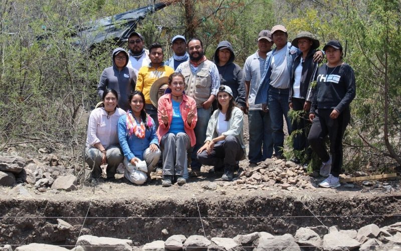 Verónica Pérez Rodríguez poses with members of the Cerro Jazmin Archaeological Project crew in Oaxaca, Mexico, in front of an archaeological excavation site populated with trees, dirt and rocks.