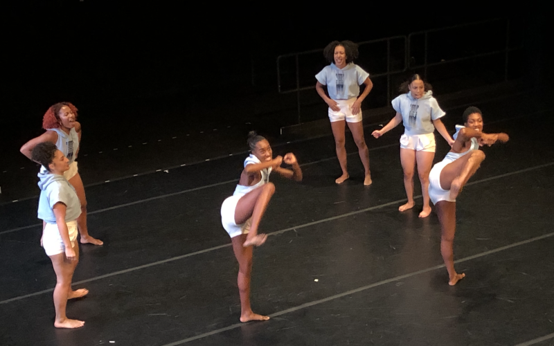 Six women in sweatshirts and shorts appear on a dark stage in varying dance poses, including knee-high kicks.