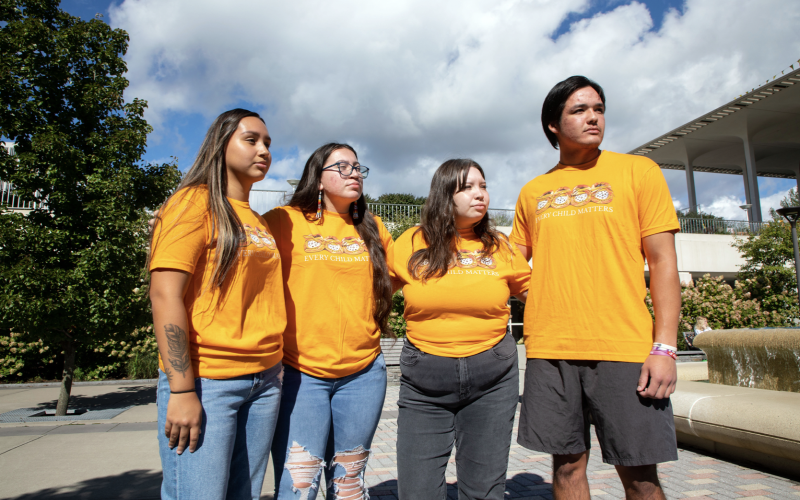 Four students in orange shirts that read "Every Child Matters" stare into the distance as they stand arm in arm next to a fountain on a sunny day. 