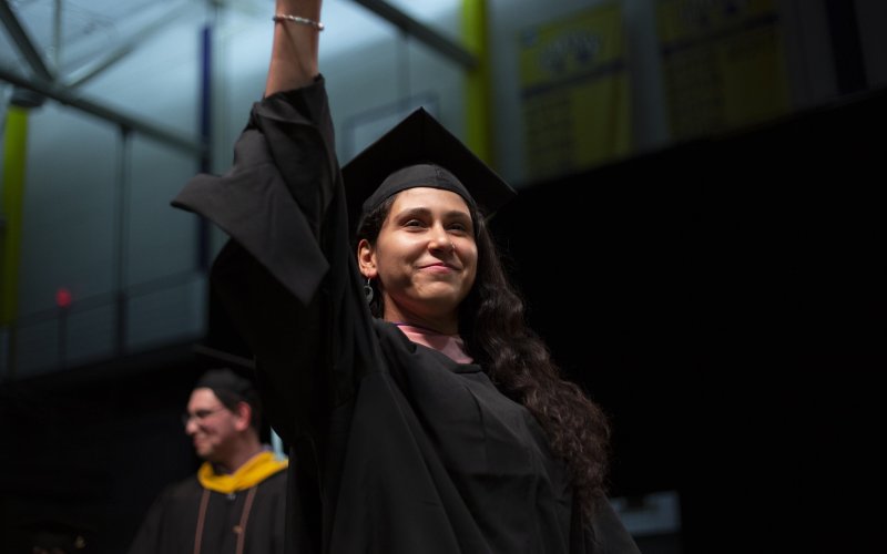 A student smiles and holds up her hand to wave as she crosses the graduation stage.