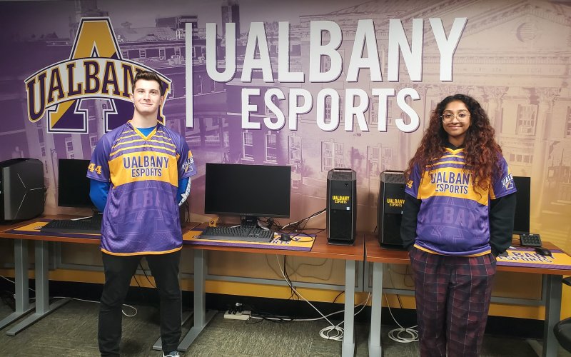 UAlbany students Dylan Tarace and Jesenia Mathew stand in front of a UAlbany eSports banner inside the competitive video gaming arena at Draper Hall.