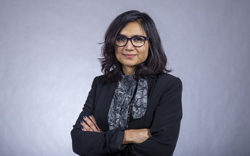 A portrait of Associate Professor Rukhsana Ahmed smiling with arms crossed in front of a plain gray background.