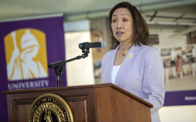 Provost Carol Kim, in a white shirt and pale purple jacket, stands at a microphone at a lecturn with the University at Albany seal on it.