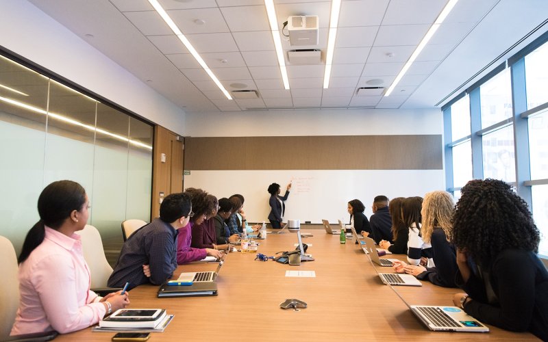 A group of professionals sit at a boardroom style table watching an African American woman at the white board giving a presentation.