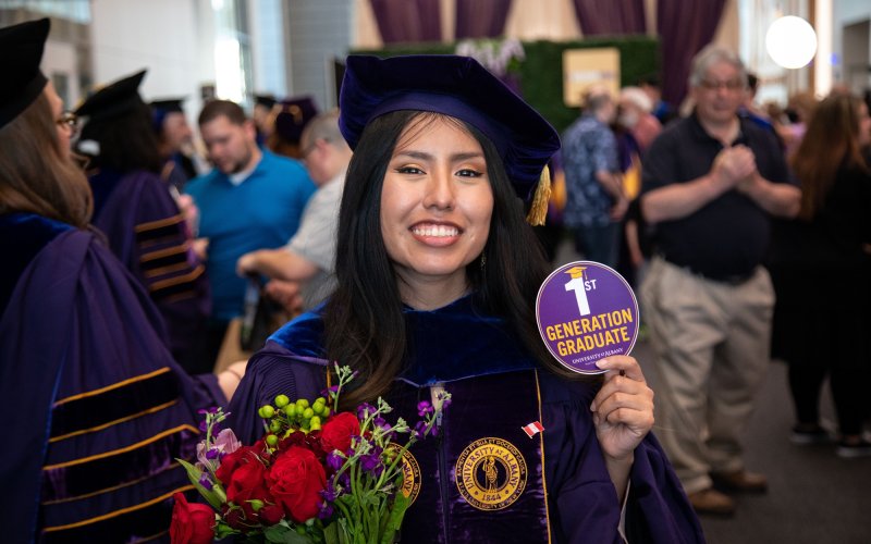 A smiling UAlbany student in cap and gown holds a bouquet of flowers and a purple sticker identifying herself as a "1st Generation Gradate."