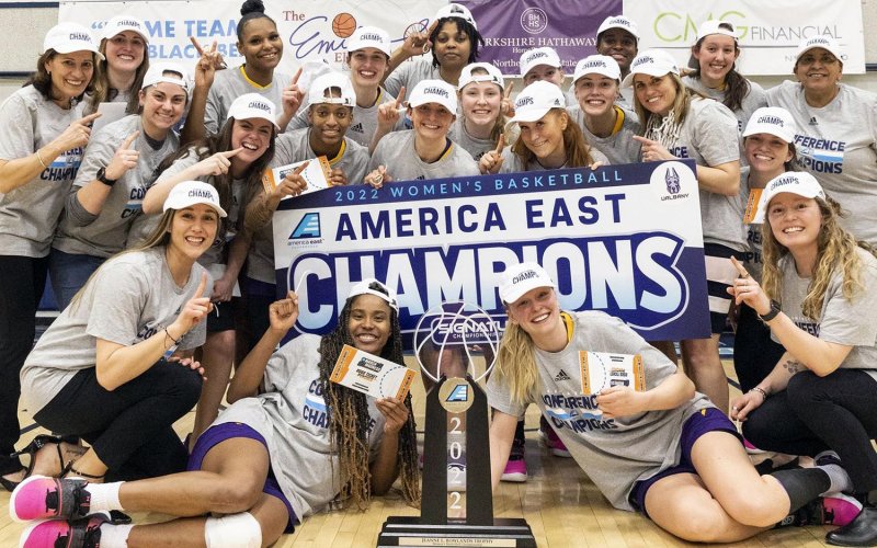 a group shot of the UAlbany women's basketball team following the America East championship in March 2022. Rows of players surround the championship trophy with a banner stating 2022 Women's basketball America East Champions.