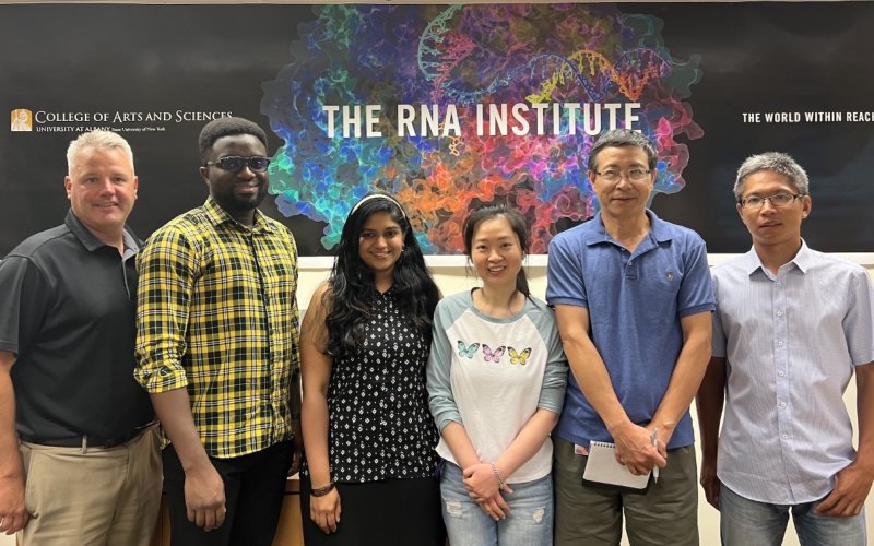 Six people stand smiling in front of an RNA Institute banner with three text headings. The headings read: "College of Arts and Sciences", "The RNA Institute" and "The world within reach". The people pictured include UAlbany faculty, staff and graduate students.