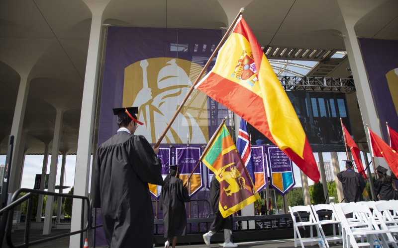 A photo of student flagbearers in graduation robes carrying various international flags onto the stage at Commencement.