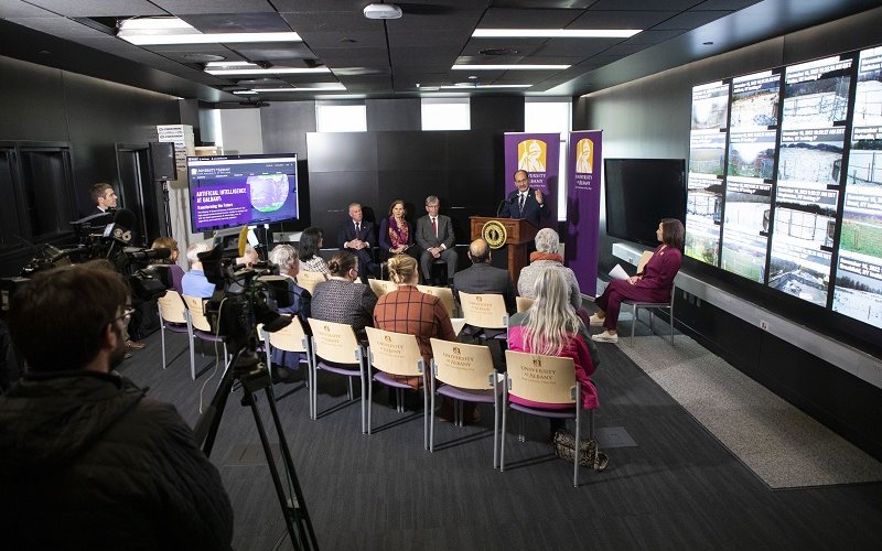 A wide-angle shot of a press conference in UAlbany's xCITE Lab. President Rodríguez stands at podium speaking flanked by two purple and gold UAlbany banners and 