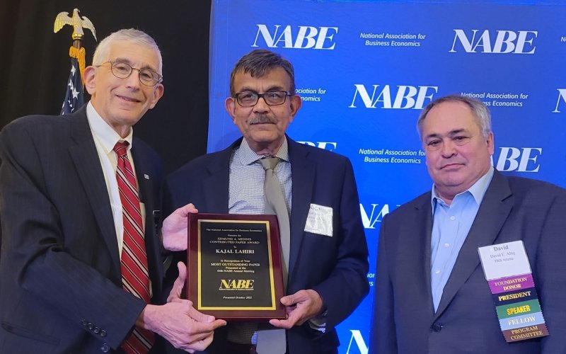 Three men in suits stand in front of a blue screen with the initials NABE printed several times. The man in the center is being handed a plaque on which his name, Kajal Lahiri, can be read.