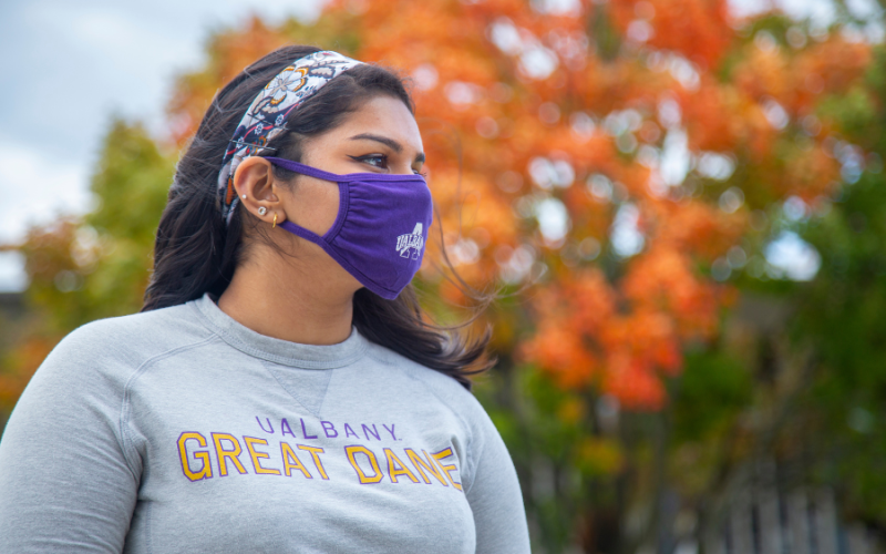 A student wearing a purple facemask in front of a tree with bright orange leaves. 