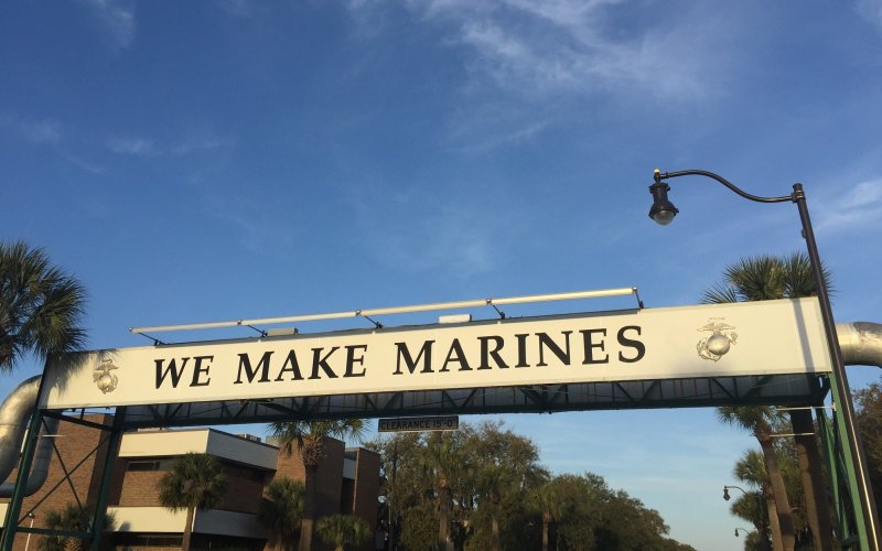 Sun illuminates a sign that reads "WE MAKE MARINES" against a backdrop of blue sky and palm trees in Parris Island, South Carolina.