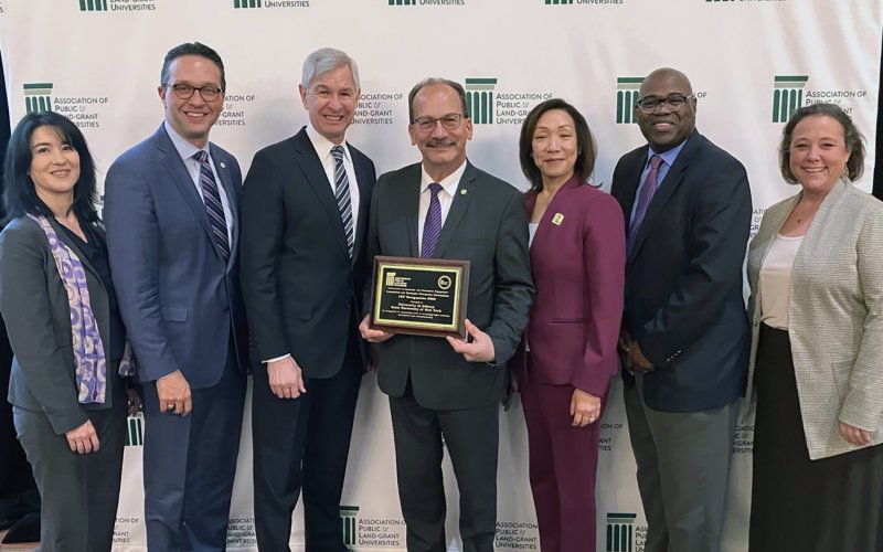 A group of seven people stand in front of a screen at the Association of Public Land-Grand Universities holding a plaque naming UAlbany as an Innovation and Economic Prosperity University.