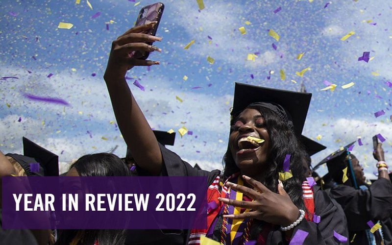A smiling UAlbany graduate holds up her smartphone to take a selfie at Commencement surrounded by a cloud of airborne purple and gold confetti, with the words "Year in Review 2022" in a purple box at the bottom left of the photo.