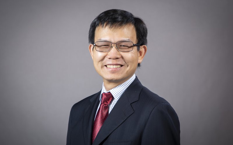 Faculty portrait of featured researcher Kai Zhang, Empire Innovation Associate Professor, School of Public Health. Professor Zhang is smiling and wearing a dark suit jacket, red silk tie, and rectangular wire rim glasses. The portrait is set against a gray backdrop. 